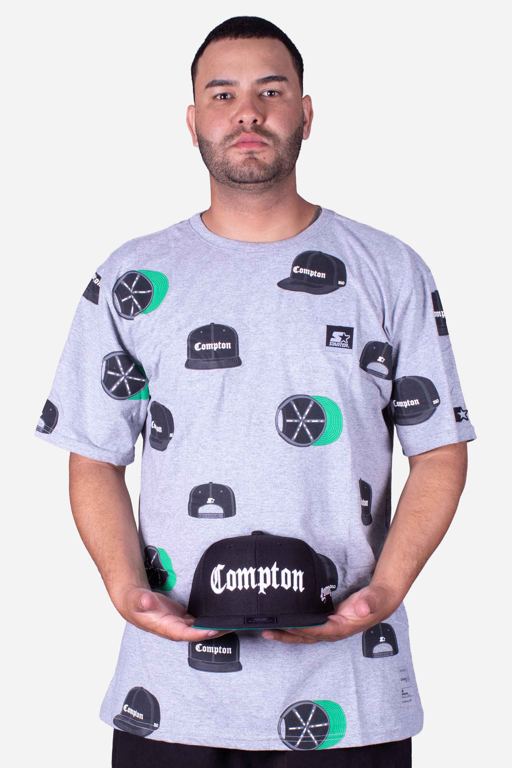 Camiseta Collab Starter x Compton Cap Cinza Mescla com estampa de bonés Compton, modelo segurando boné Compton.